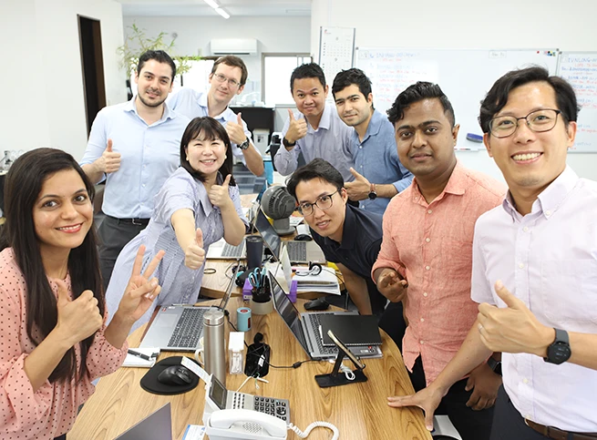 A diverse team of smiling colleagues gathered around a ta le with laptops, giving thumbs-up and peace signs in an office environment.