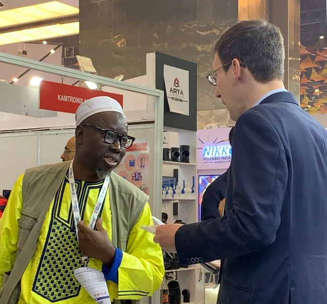 A trade show attendee in a bright yellow traditional outfit engaging in conversation with a business professional, discussing products or services at an industry event.