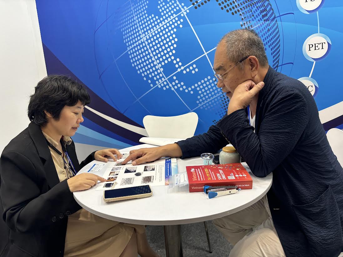 Two individuals, a client on the left and Daisaku President Mr. Kobayashi on the right discussing about products, one holding a brochure and the other engaged in conversation at a trade show booth.